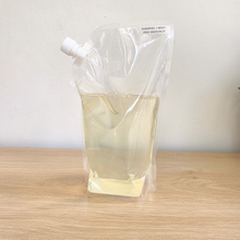 Clear refill pouch labeled "Shampoo + Body - Pink Grapefruit," filled with pale yellow liquid, resting on a wood surface.