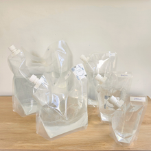 Five clear plastic flexible pouches with white caps arranged on a light wood surface against a plain off-white wall. All pouches contain water to show size when full to product weight and small handwritten labels are attached to all bags showing the size.
