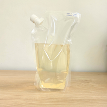 A clear plastic flexible pouch with a white screw cap, 3/4 filled (= to 1000g) with a pale yellow liquid, standing on a light wood surface against a plain off-white wall.